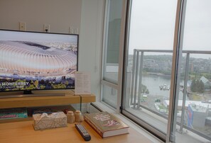 Ein Fernseher zeigt ein Stadion, ein Buch mit dem Titel 'The Ultimate Sneaker Book' und eine Fernbedienung auf einem Holztisch neben einem Fenster mit Blick auf die Stadt.