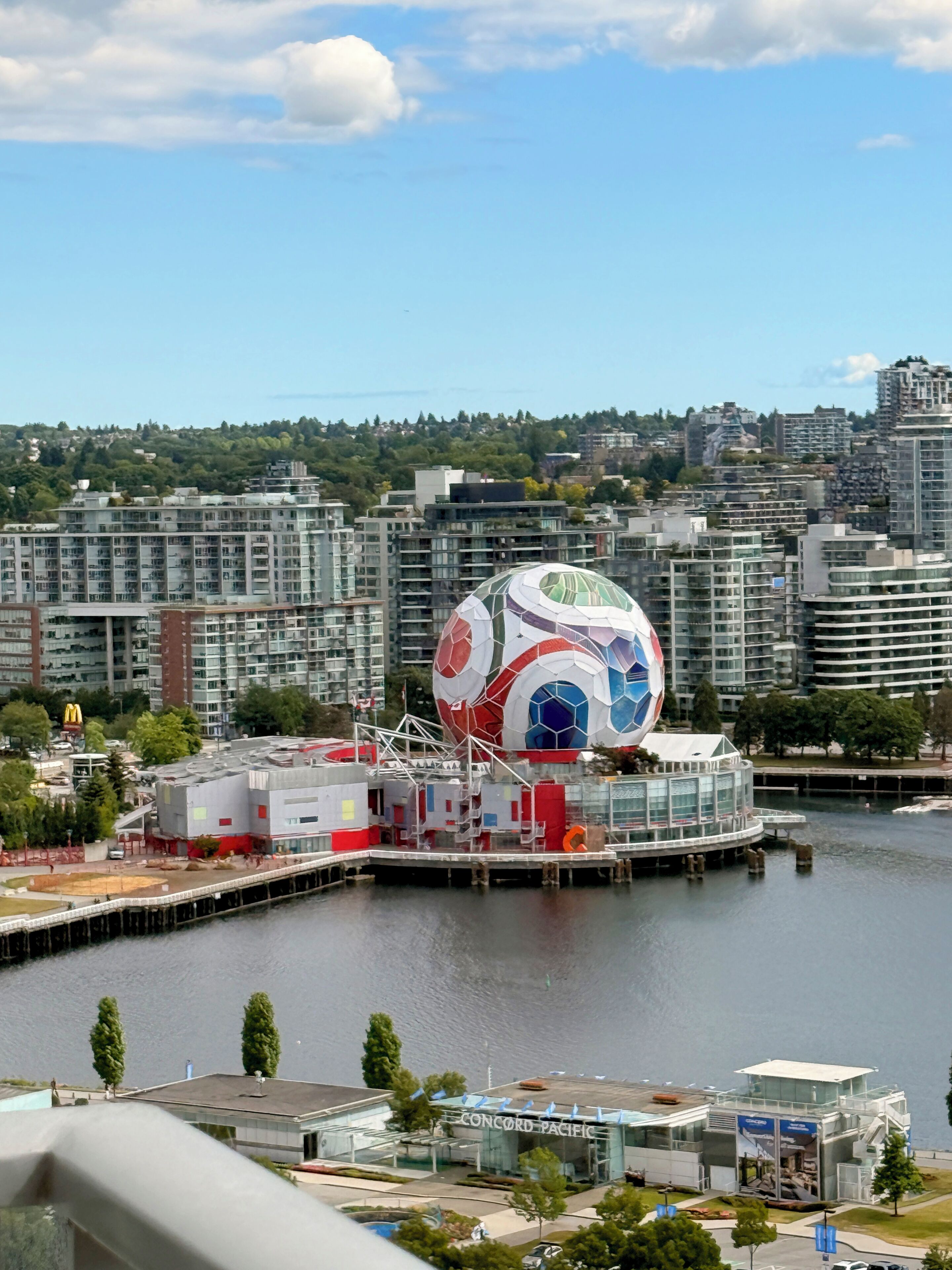 A waterfront view with a distinctive soccer ball-shaped building, a modern glass structure, and a backdrop of urban high-rise buildings.