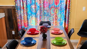 Una mesa de comedor dispuesta con platos coloridos y un jarrón de flores, con una cortina de colores al fondo.