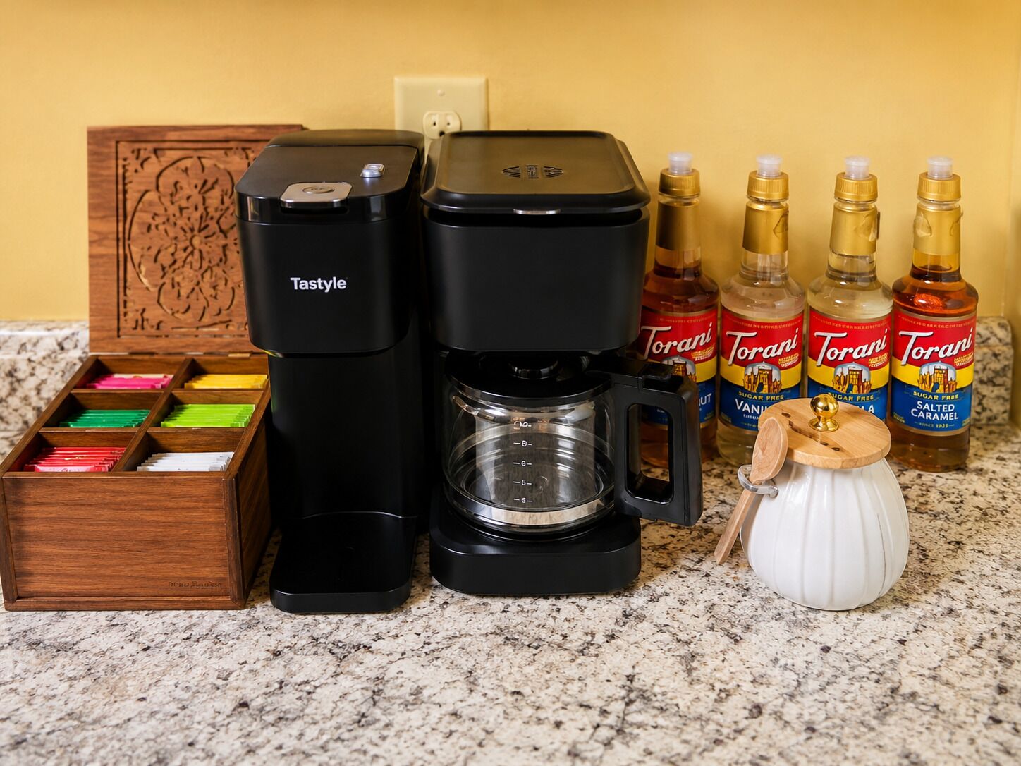 A coffee maker, Torani syrup bottles, a spice jar, and a wooden box with colored items.