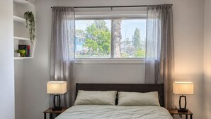 Quarto com uma cama grande, duas luminárias de cabeceira, uma janela com cortinas, uma estante com plantas e uma luminária no teto.