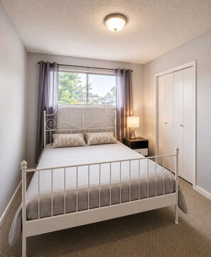 A single bed with a metal frame, a bedside table with a lamp, a window with curtains, and a door.