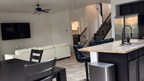 A modern kitchen with a central island, a dining area with a table and chairs, a flat-screen TV mounted on the wall, and a staircase leading to an upper level.