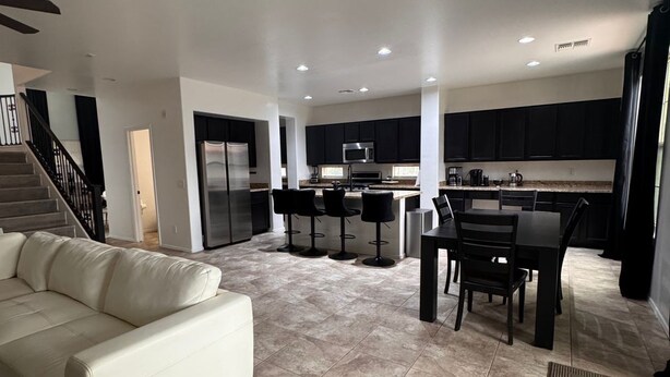 A modern living room with a sofa, a dining area with chairs, a kitchen with dark cabinets and appliances, and a staircase.