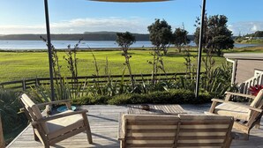 A wooden deck with two lounge chairs and a table, overlooking a grassy field and a body of water. - A restorative stay where the mighty Waikato awa meets the Tasman Sea (Port Waikato)