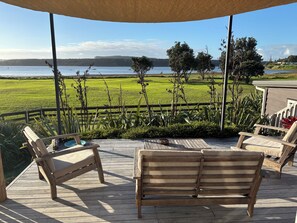 A wooden deck with two lounge chairs and a table, overlooking a grassy field and a body of water. - A restorative stay where the mighty Waikato awa meets the Tasman Sea (Port Waikato)