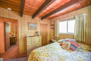A room with wooden walls, a bed with floral bedding, a dresser, a desk, and a window with curtains.