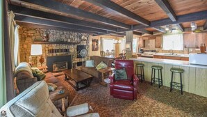 Un intérieur rustique avec une cheminée en pierre, des poutres apparentes en bois au plafond, un coin repas et une cuisine équipée de tabourets de bar.