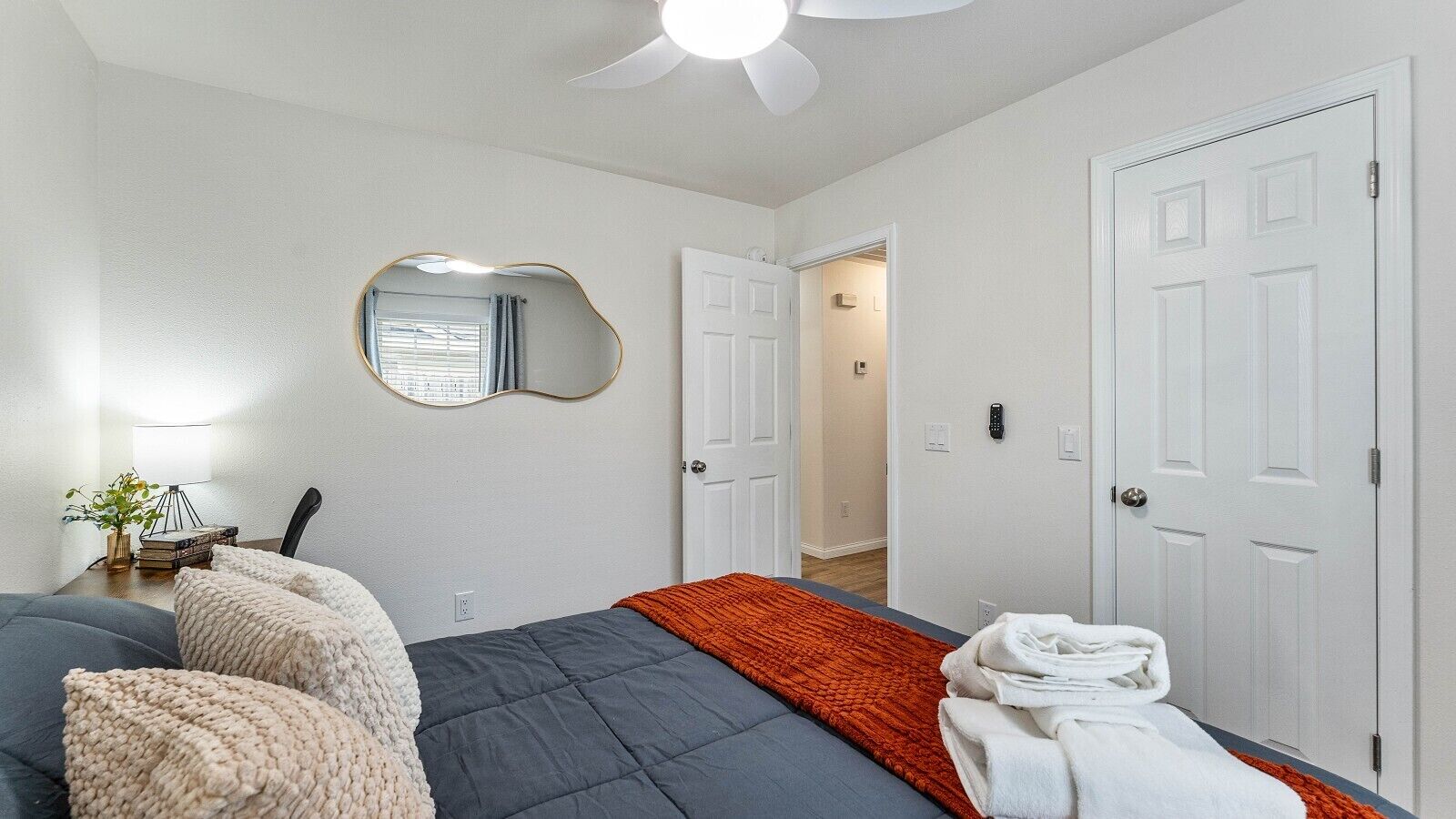 A bedroom with a bed, a mirror, a bedside table with a lamp, and a door leading to another room.