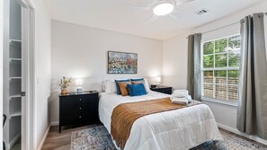 Um quarto com cama, mesas de cabeceira, um quadro, uma janela com cortinas e um ventilador de teto.