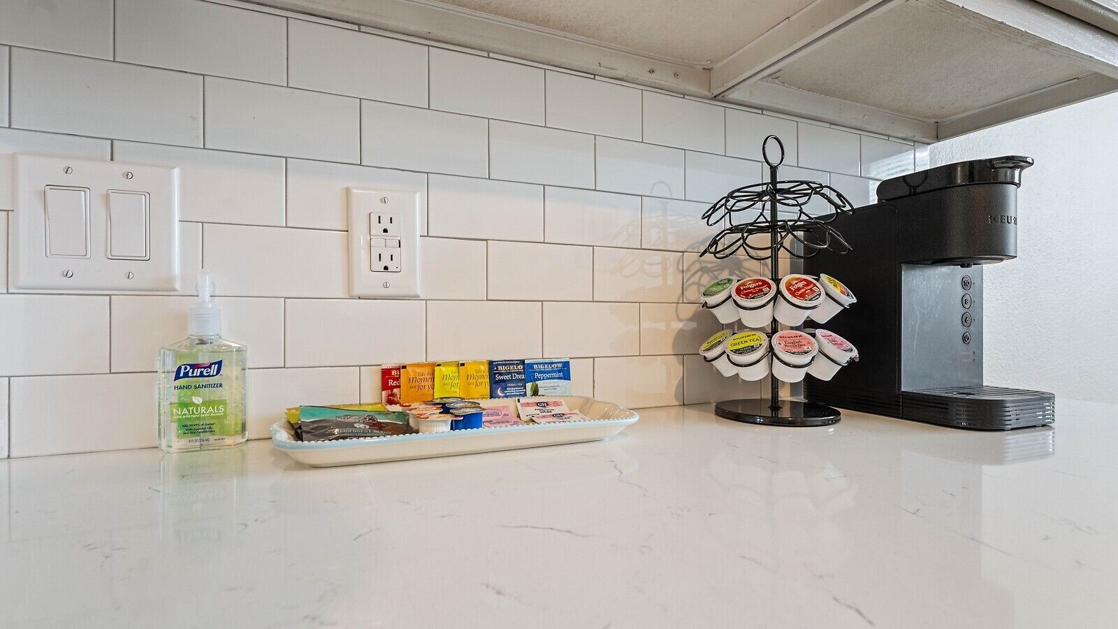 Una encimera de cocina con dispensador de gel hidroalcohólico, una bandeja de aperitivos, una máquina de café y un dispensador de té.