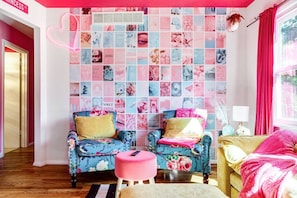 Ein Zimmer mit Wandcollage, zwei blumige Sessel, ein pinkfarbener Hocker und ein Bett mit rosa Decke.