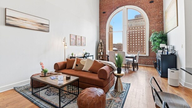 Un salon moderne avec un canapé marron, une table basse et un pouf, un coin repas avec des chaises et une table, un coin cuisine avec une cuisinière et des placards, et une grande fenêtre en arc de cercle offrant une vue sur la ville.