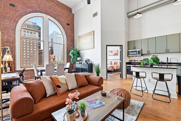 A modern apartment with a large window, a kitchen, a living area with a sofa, and a dining area.