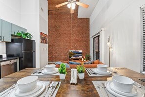 Une cuisine moderne avec un mur en briques, un ventilateur de plafond en bois et un coin repas doté d’une longue table dressée pour quatre personnes.