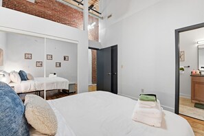 Un dormitorio moderno con una cama grande, una cama más pequeña, un muro de ladrillo y una puerta que da a un baño.