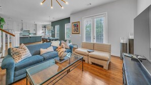 A spacious living area with a blue sofa, beige sofa, and a glass coffee table. There is a kitchen in the background with white cabinets and a dining area. - THE PARKER, A Modern Retreat Near Everything: 3 BR, 3.5 bathroom house Sleeps 8 (Houston)