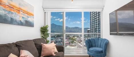 Ein modernes Wohnzimmer mit einer Couch, einem blauen Sessel, einem großen Fenster mit Stadtblick und einer Deckenleuchte.