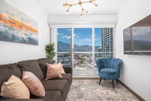 Ein modernes Wohnzimmer mit einer Couch, einem blauen Sessel, einem großen Fenster mit Stadtblick und einer Deckenleuchte.