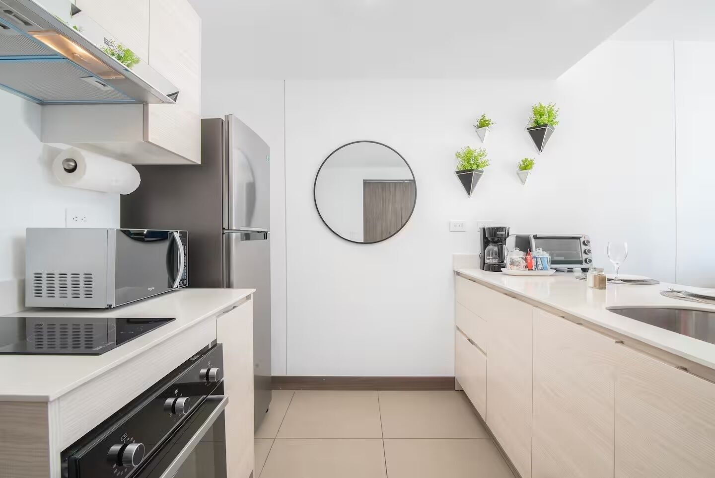 Cozinha moderna com forno embutido, micro-ondas e geladeira, espelho redondo e plantas em vasos na parede.
