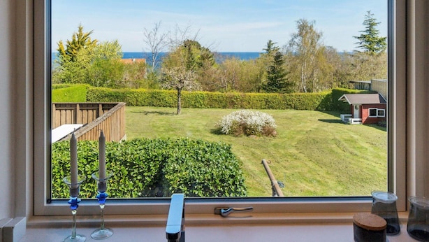 A window with a view of a well-maintained garden, a wooden deck, and a small building in the background. - 5 Star Holiday Home in Allinge (Allinge)