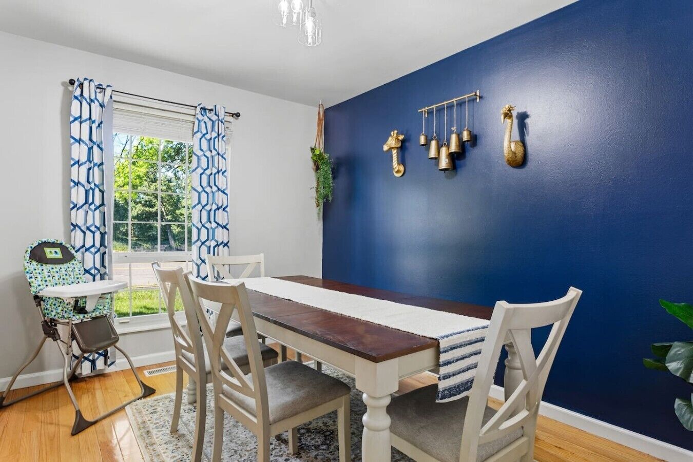 Comedor con mesa de madera, sillas blancas, pared azul y ventana con cortinas estampadas.