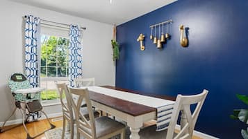 Comedor con mesa de madera, sillas blancas, pared azul y ventana con cortinas estampadas.