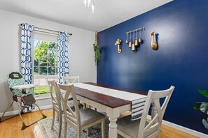 Comedor con mesa de madera, sillas blancas, pared azul y ventana con cortinas estampadas.