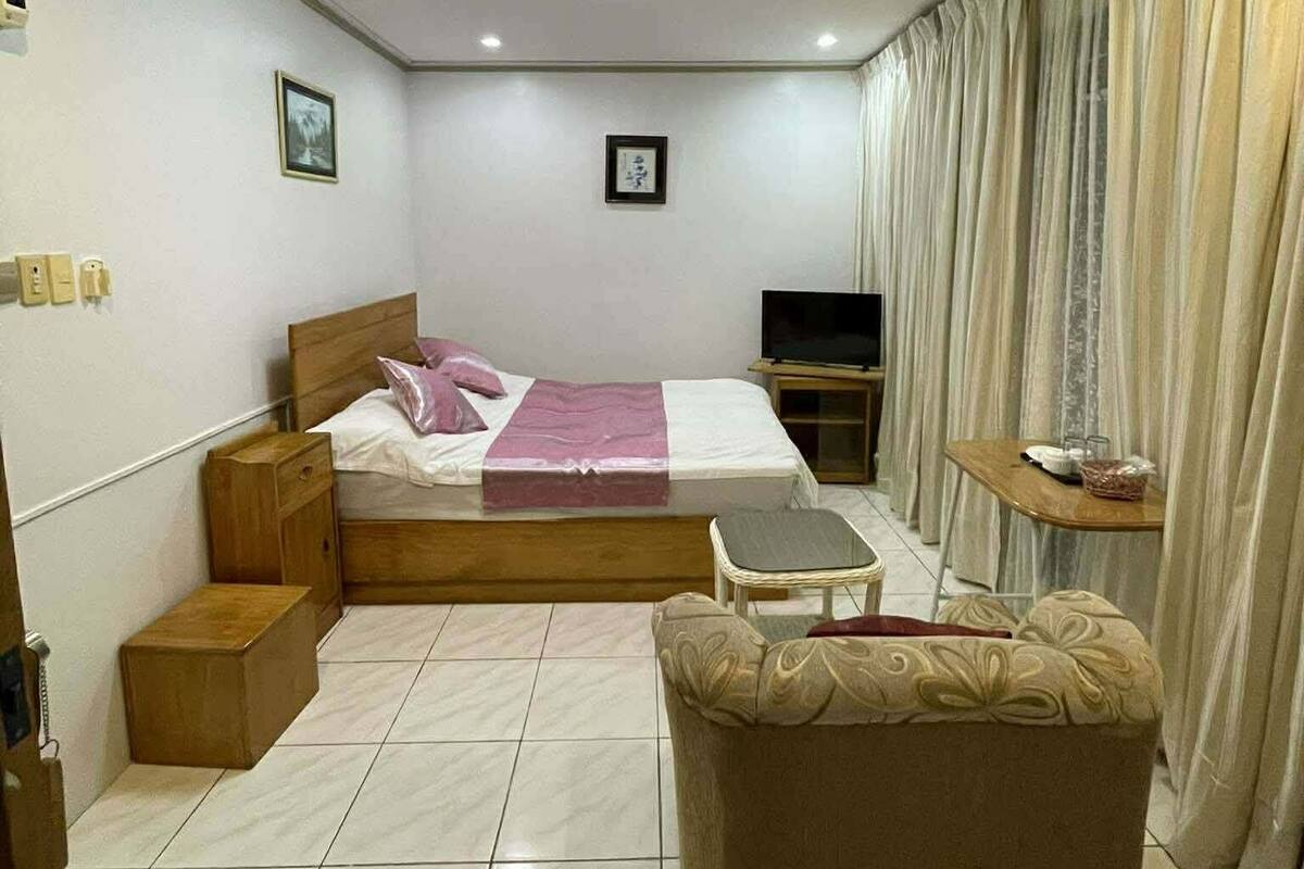 A bedroom with a bed, a TV, a small table, a sofa, curtains, and a ceiling light.