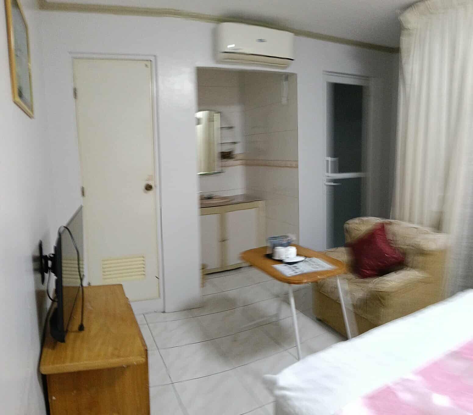 Hotellihuone, jossa on sänky, pieni pöytä, jossa on kuppi, tuoli, vaatekaappi ja ilmastointilaite.
