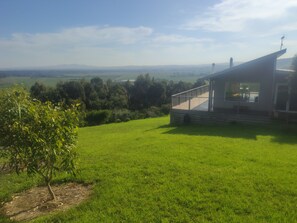 A modern house with a large balcony overlooking a grassy area and distant hills.