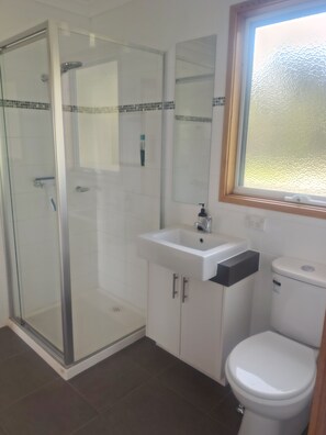 Ein Badezimmer mit einer gläsernen Duschkabine, einem weißen Waschbecken mit schwarzem Seifenspender und einer weißen Toilette.