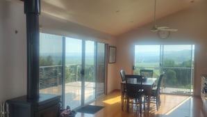 A spacious room with a wood-burning stove, large sliding glass doors, a dining table with chairs, and a ceiling fan. - Whole House in Toora on 2 ha. elevated with incredible views to Wilsons Prom (Toora)