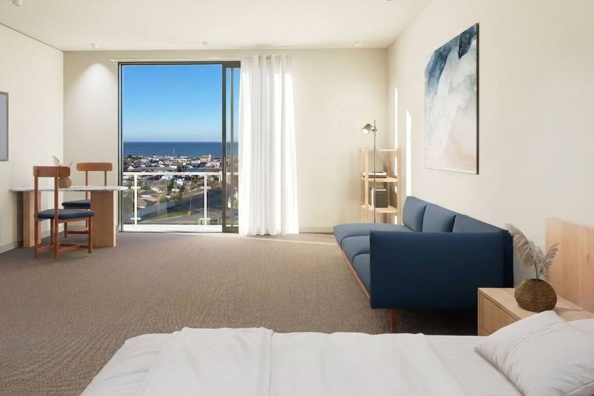 Habitación de hotel con cama, un sofá azul, un escritorio con silla, un ventanal con vistas a la ciudad y un cuadro en la pared.