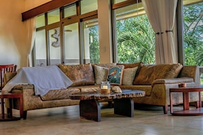 Un salón acogedor con un sofá de cuero marrón, una mesa de centro de madera y vistas a exuberante vegetación a través de grandes ventanales.