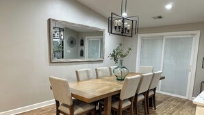 Un coin repas avec une table et des chaises en bois, un luminaire suspendu, un miroir et une fenêtre avec des stores.
