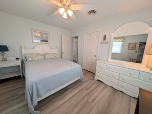 Quarto com cama, penteadeira, mesa de cabeceira, ventilador de teto e uma porta.