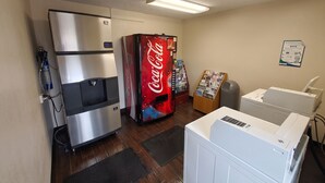 Une pièce équipée d’un réfrigérateur, d’un distributeur de Coca-Cola, d’un tableau d’affichage, d’une poubelle et d’un sèche-linge.