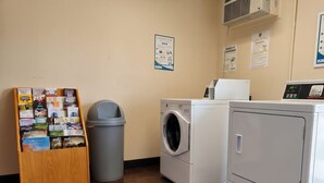 Un espace buanderie comprenant un présentoir en bois rempli de brochures, une poubelle grise, un lave-linge à chargement frontal et un sèche-linge superposé.
