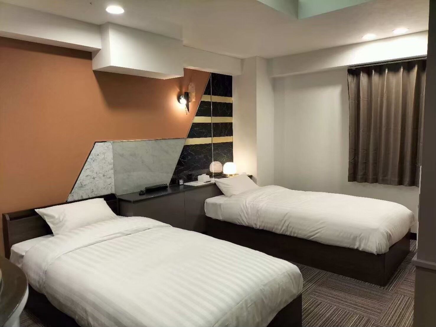 A hotel room with two beds, a marble accent wall, a large mirror, and a window with curtains.