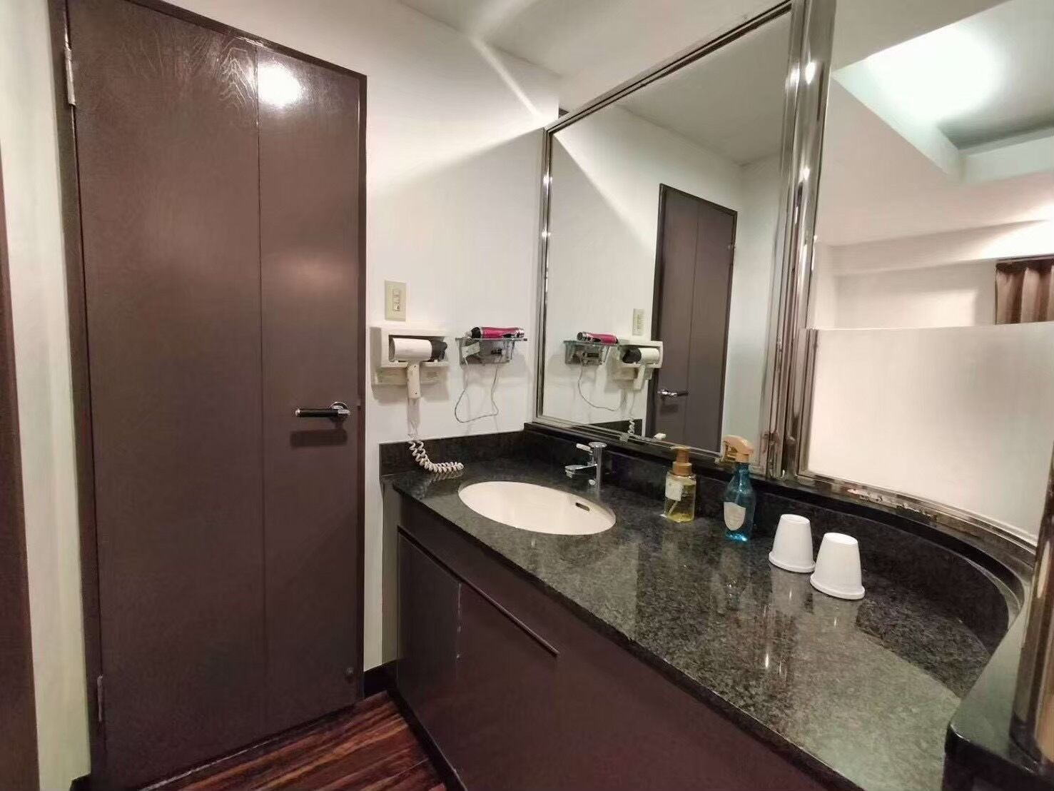 Un baño de hotel con una puerta de madera oscura, un lavamanos blanco, un espejo grande y un perchero para toallas.