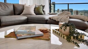 Una sala de estar moderna con un sofá de cuero, una mesa de centro con libros y una planta, y vistas a la ciudad a través de grandes ventanales.