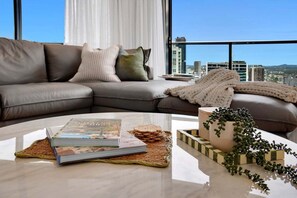 Ein modernes Wohnzimmer mit einer Ledersofa, einem Couchtisch mit Büchern und einer Pflanze, und Blick auf die Stadt durch große Fenster.