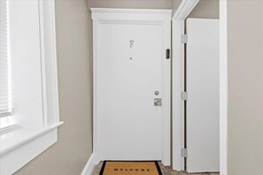 Una puerta blanca con cerradura electrónica, un felpudo con la palabra 'WELCOME' y una ventana con persianas.