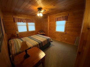 A log cabin bedroom with a bed, a chair, a table, and two windows with curtains.