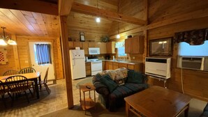 Interior acollidor d'una cabana, amb zona de dinar, sofà, taula petita, cuina amb electrodomèstics bàsics i televisió penjada.