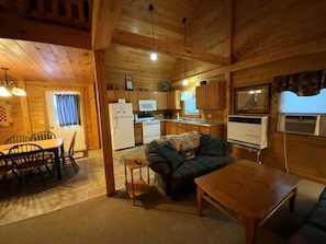 Ein gemütliches Blockhaus-Interieur mit Essbereich, einer Couch, einem kleinen Tisch, einer Küche mit Küchengeräten und einem Wand-Fernseher.