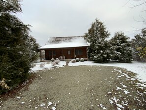 A wooden cabin with a covered porch, surrounded by snow-covered trees and a gravel driveway.