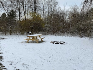 A snow-covered picnic table in a forested area with bare trees.