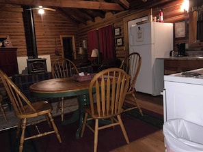 A wooden cabin interior with a dining table, chairs, a kitchen area, and a fireplace.